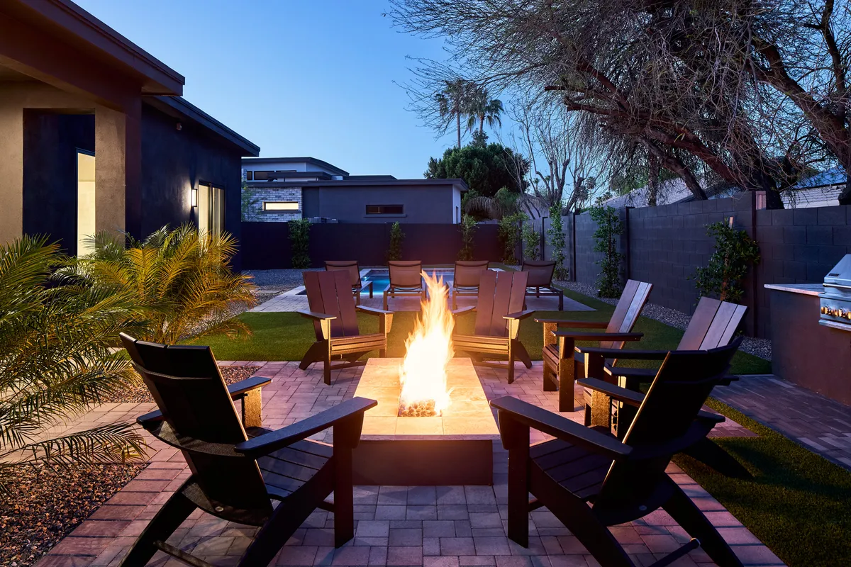 A cozy backyard setting with a glowing fire pit surrounded by comfortable chairs, a sparkling swimming pool, and lush landscaping. The atmosphere is warm and communal, perfect for gathering with friends in the evening.