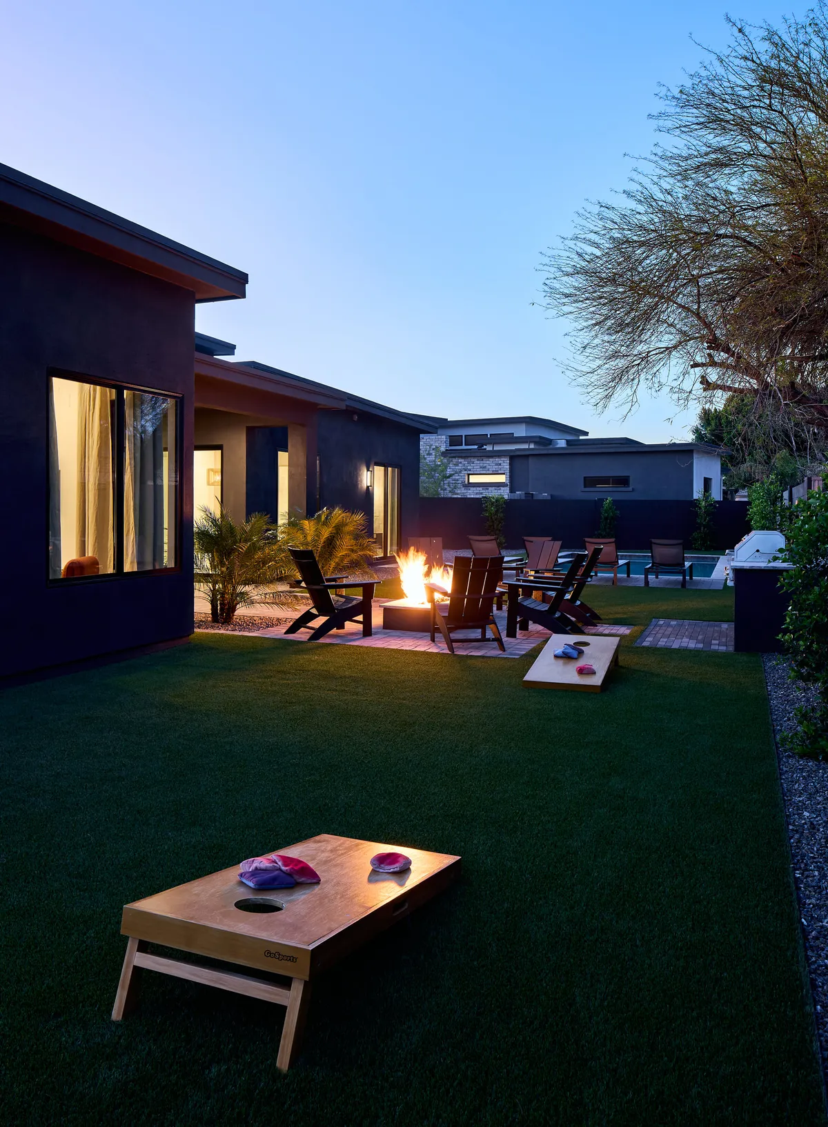 The image features a lush lawn with cornhole boards set up, a fire pit circled by Adirondack chairs, and an overall relaxed, playful atmosphere. It evokes family fun, friendly games, and cozy evenings under the stars.