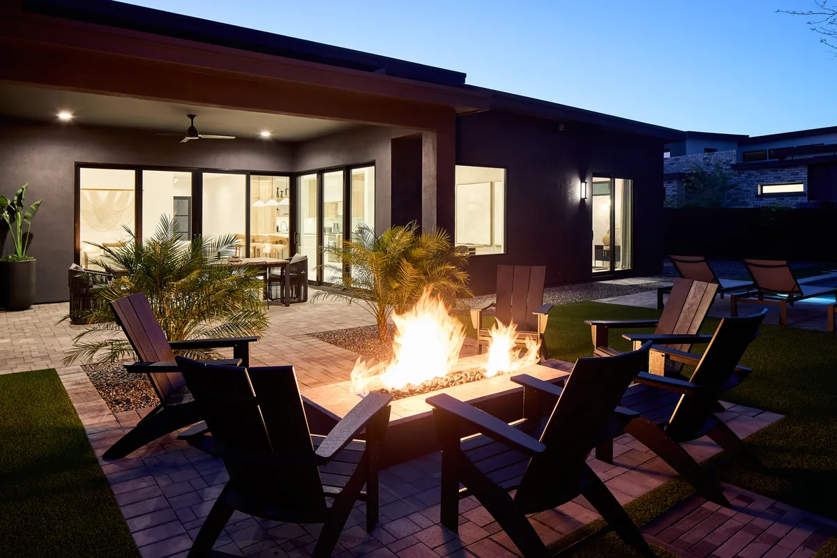 The outdoor entertaining area at Tulum House boasts a glowing fire pit, comfortable seating, lush palm plants, and expansive windows that connect the indoor and outdoor spaces. The setting is perfect for evening gatherings or informal business chats under the Arizona sky.