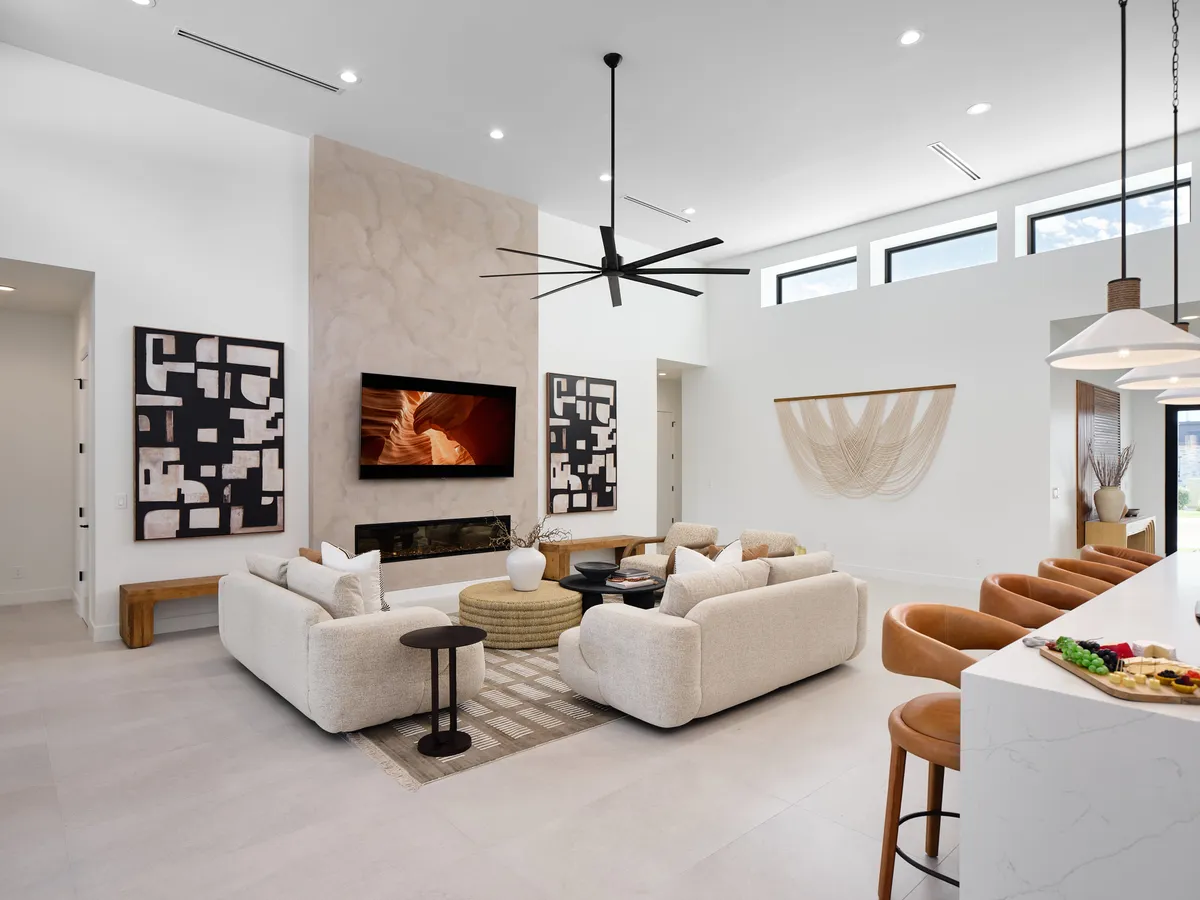 A spacious open-plan living area featuring a large sectional sofa, modern art on the walls, and a sleek fireplace with a TV mounted above. The room flows seamlessly into a stylish kitchen island visible in the background, and natural light fills the space, highlighting designer furnishings and plush seating ideal for gatherings.